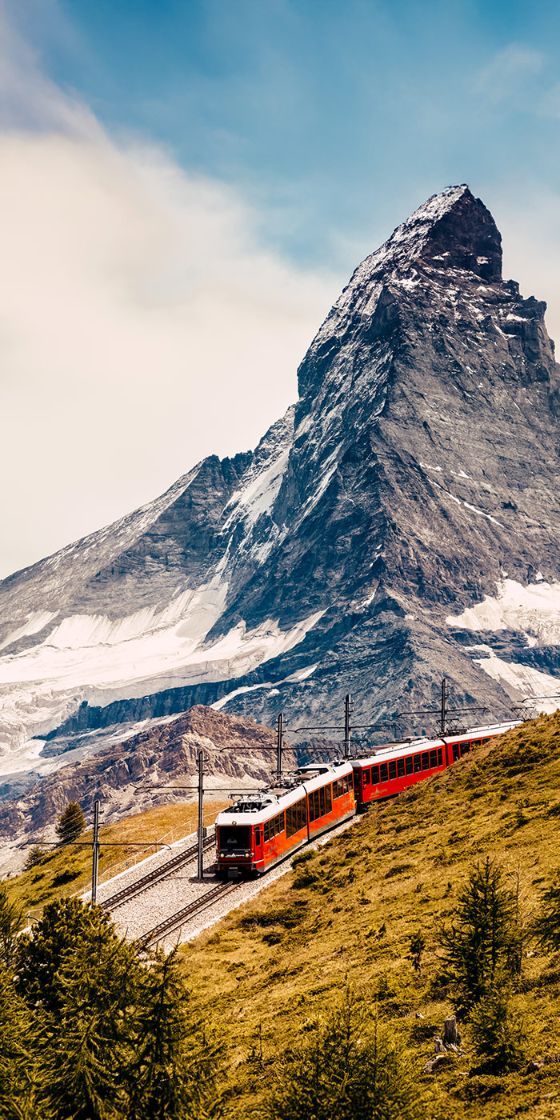 Eine rote Bergbahn fährt auf einer Trasse über einen grasbewachsenen Bergkamm vor der Kulisse eines schneebedeckten Berggipfels.