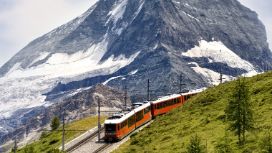 Eine rote Bergbahn fährt auf einer Trasse über einen grasbewachsenen Bergkamm vor der Kulisse eines schneebedeckten Berggipfels.