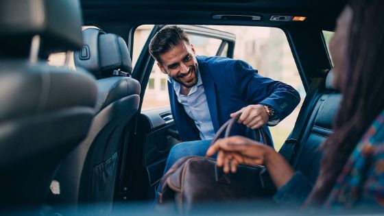 Blick einer Frau von der Rückbank eines SUVs auf einen lächelnden Mann im blauen Jackett mit Ledertasche beim Einsteigen