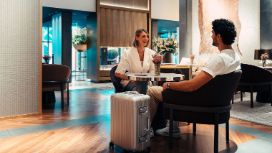 Eine Frau und ein Mann sitzen in einer eleganten Hotel-Lobby an einem Tisch, vor ihnen steht ein schicker Rollkoffer.
