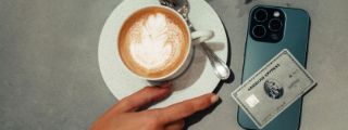 Eine Hand greift nach einer Tasse Cappuccino, die auf einer grauen Tischplatte steht, daneben liegt ein Smartphone mit einer Kreditkarte von American Express.