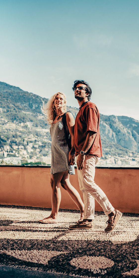 Ein Paar in sommerlicher Kleidung steht händchenhaltend auf einer Plattform mit gelber Mauer vor einer mediterranen Berglandschaft bei Sonnenschein.