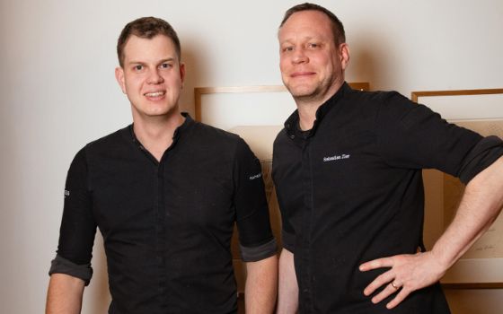 Vor einer hellen Wand stehen zwei Männer, sie haben kurze, dunkle Haare und beide tragen ein schwarzes Hemd. Auf der linken Seite steht Richard Schmidtkonz, der Head Chef und rechts daneben Sebastian Zier, Executive Chef. 