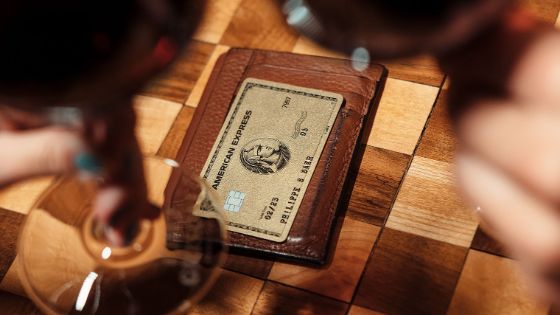 Eine goldene Kreditkarte von American Express auf einem braunen Lederetui aus auf einem gemusterten Holztisch, gerahmt von zwei gefüllten Rotweingläsern