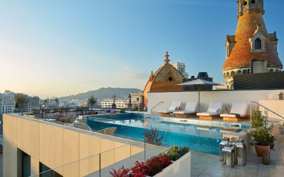 Eine luxuriöse Hotelterrasse mit Pool und Sonnenliegen über den Dächern Barcelonas unter blauem Himmel im warmen Abendlicht