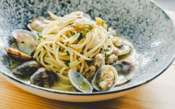 Spaghetti und Muscheln sind mit Kräutern auf einem hellgrauen Teller angerichtet. 