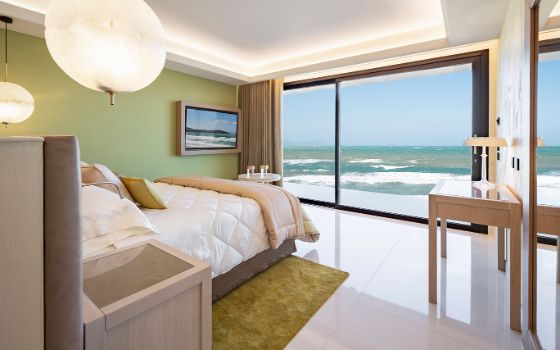 Minimalistisches Hotelzimmer in hellen Farbtönen mit Doppelbett und Blick aufs Meer durch ein bodentiefes Panoramafenster.