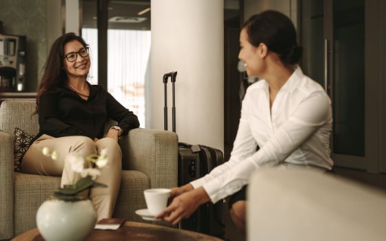 Eine Frau sitzt in einem Sessel und lächelt eine Kellnerin an, die ihr Kaffee serviert, neben ihr steht ein Rollkoffer.