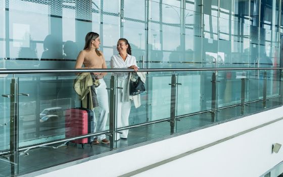 Zwei gut gelaunte Frauen mit Handgepäck stehen auf einer Empore vor einer Glaswand in einem Flughafenterminal.