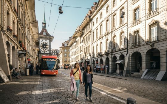 Strassenszene in der Berner Altstadt mit Blick auf die Zytglogge, rotem Bus Richtung Zentrum Paul Klee und Fussgängerinnen unter den historischen Lauben.
