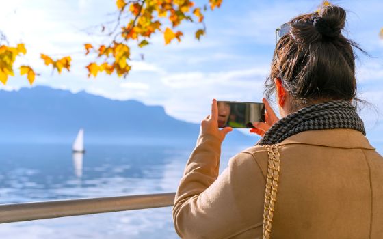 Frau fotografiert mit dem Smartphone den Genfersee mit Segelboot, Herbstlaub und Alpenkulisse im Hintergrund.