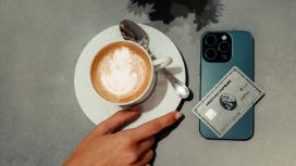 Eine Hand greift nach einer Tasse Cappuccino, die auf einer grauen Tischplatte steht, daneben liegt ein Smartphone mit einer Kreditkarte von American Express.