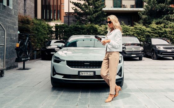 Eine elegant gekleidete Frau mit Sonnenbrille geht vor einem weissen, geparkten Auto auf einem Hotelparkplatz.