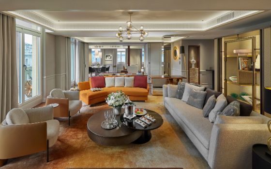 Helle, luxuriöse Hotelsuite mit Panoramafenstern, Teppichboden und verschiedenen Sitzpolstern um einen runden Couchtisch gruppiert, auf dem Champagner und kleine Speisen stehen; im Hintergrund ein Esszimmer.
