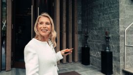 Frau in edler weisser Jacke steht vor einem Eingang, blickt hinter sich und lächelt.