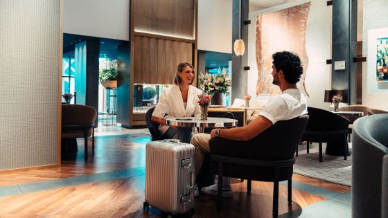 Eine gut gelaunte Frau und ein Mann sitzen an einem Tisch in einem stilvollen Hotelfoyer, neben ihnen steht ein hochwertiger Aluminiumkoffer.