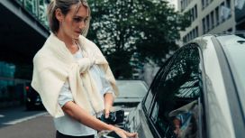Eine moderne, elegant gekleidete Frau steht an einem geparkten Auto auf einer Strasse in urbaner Umgebung, in einer Hand hält sie ihr Smartphone und den Autoschlüssel.
