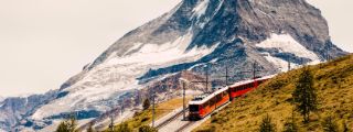 Eine rote Bergbahn fährt auf einer Trasse über einen grasbewachsenen Bergkamm vor der Kulisse eines schneebedeckten Berggipfels.