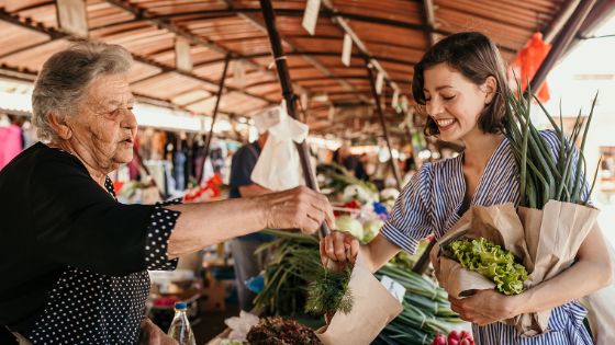 Eine Frau mit Blumen und Gemüse im Arm kauft beim Marktstand einer älteren Dame Kräuter ein.