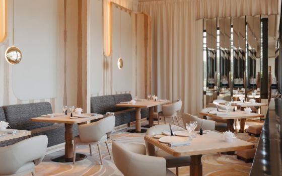 Im Inneren des Restaurants stehen hellgraue Sofas an den Wänden, davor quadratische Holztische mit beigen Stühlen. 