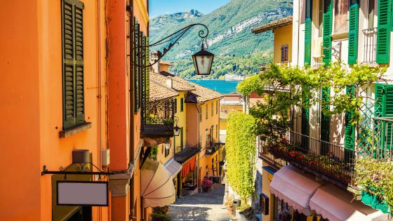 Gasse mit Treppenstufen in einem mediterranen Ort mit bunten Hausfassaden, im Hintergrund ein See vor Bergpanorama