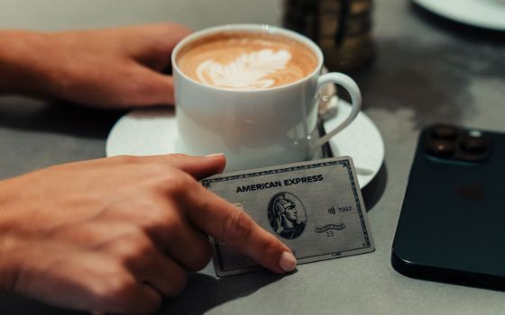 Eine silberne Kreditkarte von American Express in einer Hand neben einem Smartphone und einer gefüllten Cappuccinotasse auf einem Tisch.